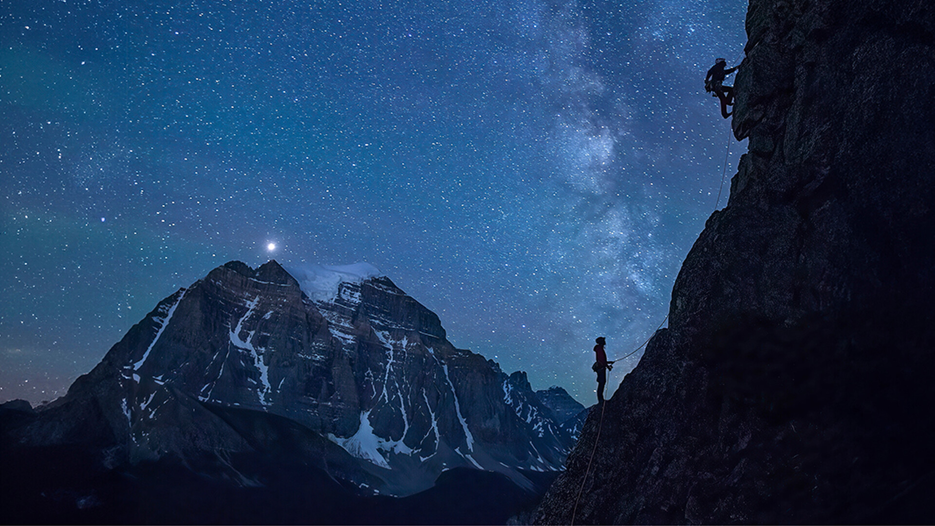Climber on a steep, overhanging rock face glowing orange, with a full moon rising over jagged rock spires against a blue sky in the background.