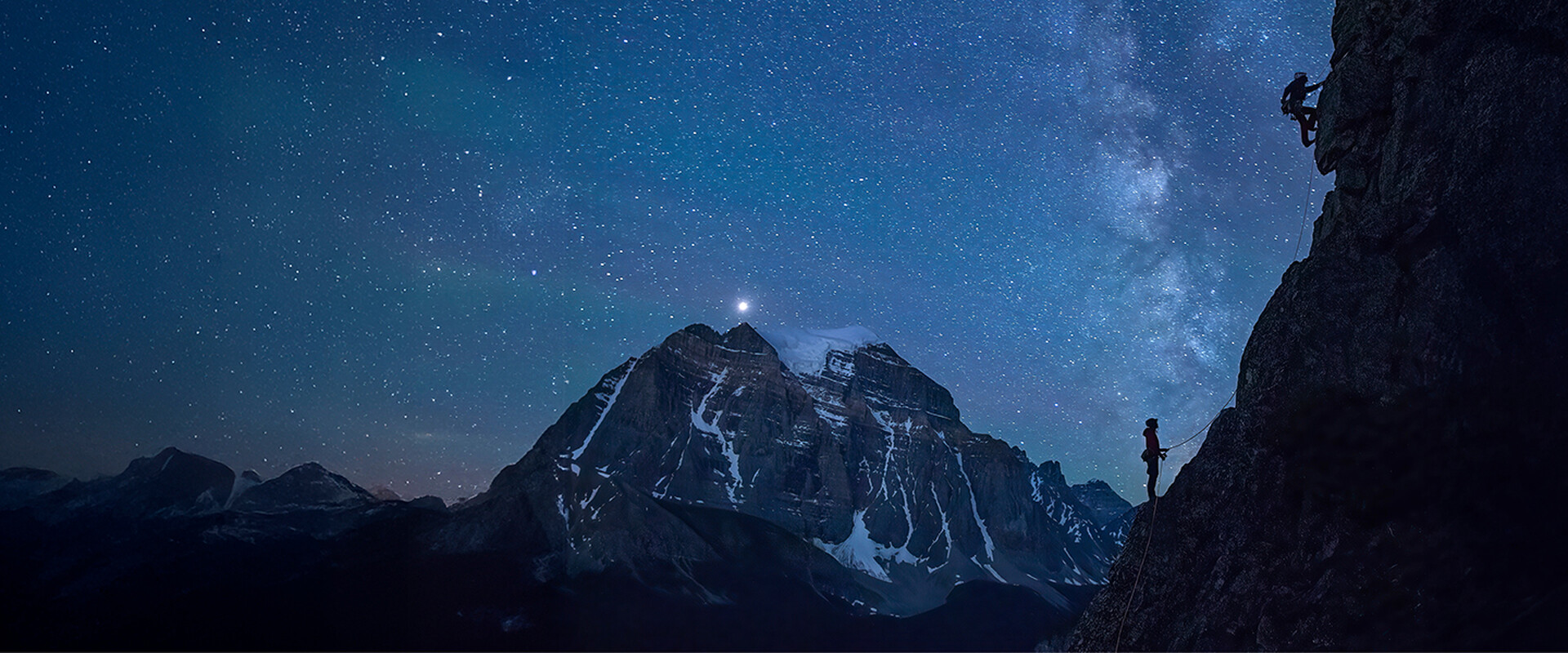 Climber on a steep, overhanging rock face glowing orange, with a full moon rising over jagged rock spires against a blue sky in the background.