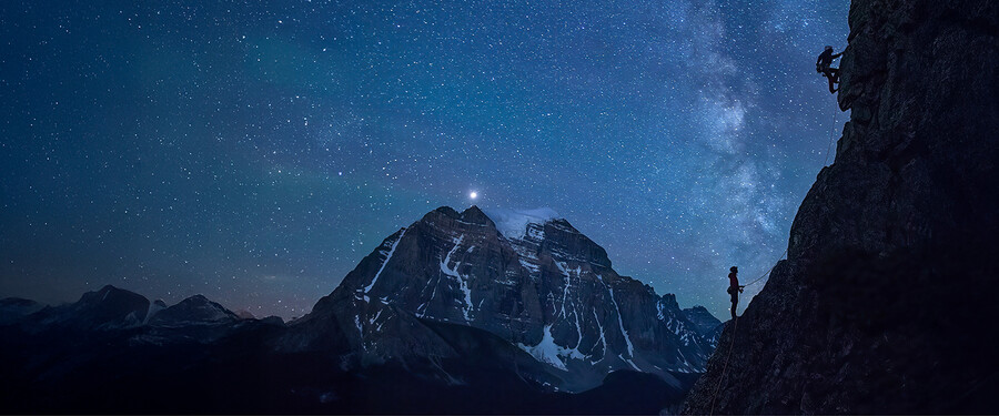 Climber on a steep, overhanging rock face glowing orange, with a full moon rising over jagged rock spires against a blue sky in the background.